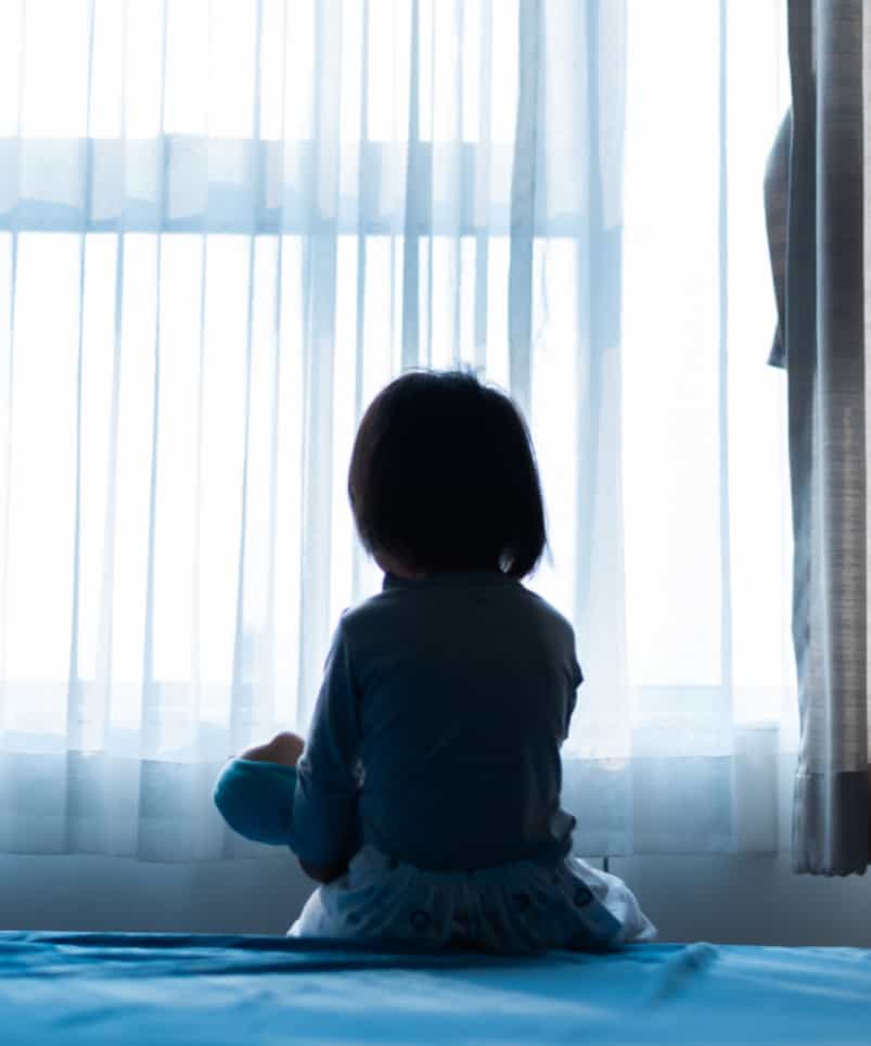 Small child sitting on a bed looking out a bright window curtain