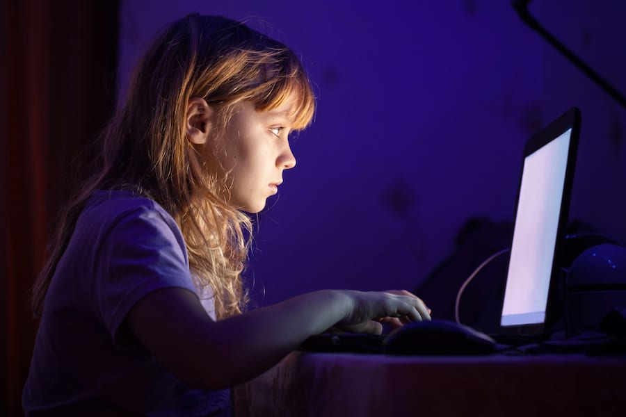 A young blond girl works on a laptop in a dark room with a glowing screen