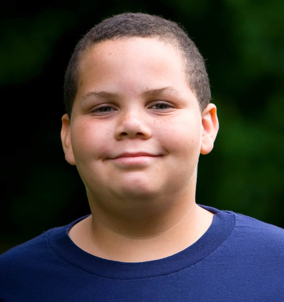 A young latino boy stands outdoors in a blue shirt, smiling at the camera.