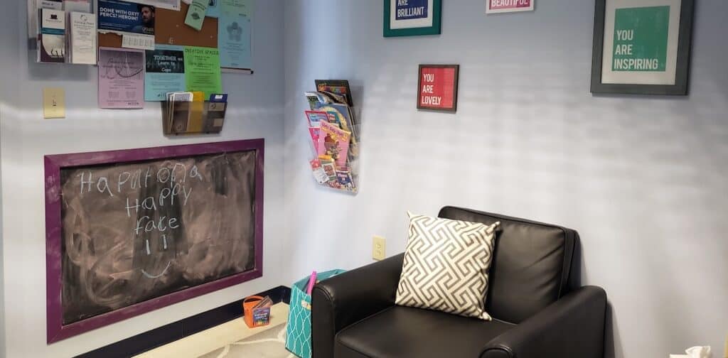 A black leather chair sits in a forensic interview room against pale walls and a chalkboard that says Put on a Happy Face.