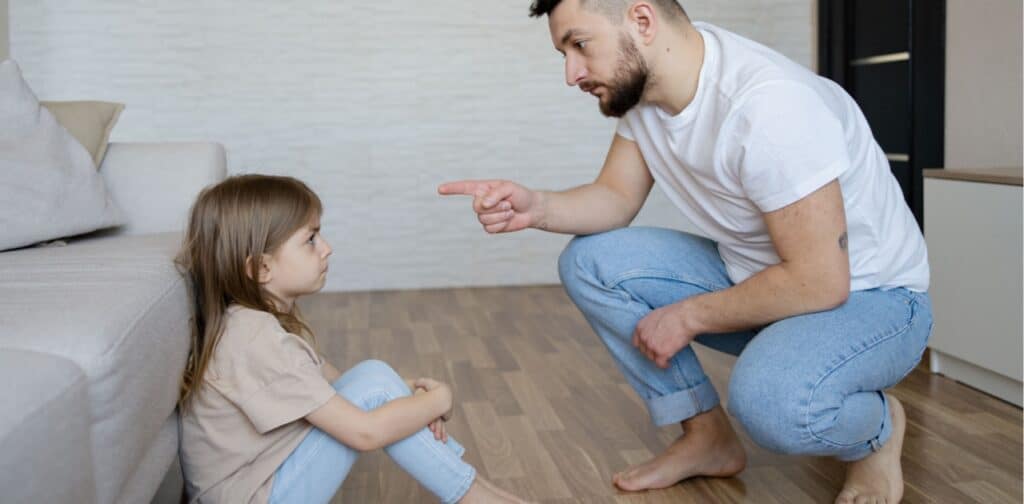 A father points at his young daughter in a living room.